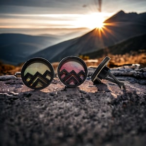 May include: Three round wooden car air fresheners with mountain designs. The air fresheners are in shades of green, pink, and black. The air fresheners are sitting on a rock surface.
