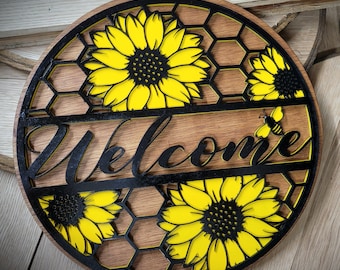 May include: A round wooden sign with a honeycomb pattern and yellow sunflowers. The sign says "Welcome" in black lettering and features a small black bee.