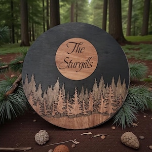 May include: A round wooden sign with a forest scene and the text "The Sturgills". The sign features a dark gray background with a laser-cut forest design and a lighter wood circle in the center.