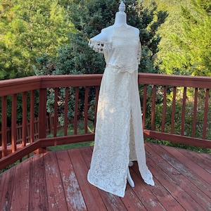 May include: A white crochet maxi dress with a halter neckline and a lace pattern. The dress has a slit up the front and is on a white mannequin. The dress is on a wooden deck with a green background.