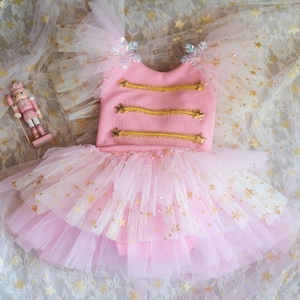 May include: A pink textured bodice dress featuring gold braided trim across the chest, ruffled tulle sleeves with snowflake accents, and a multi-layered tulle skirt adorned with gold star patterns, accompanied by a small nutcracker figurine.