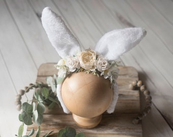 May include: White bunny ears headband with a floral crown of white and cream flowers. The headband is on a wooden mannequin head.