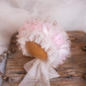 May include: A white and pink ruffled baby bonnet with a satin bow. The bonnet is made of tulle and has a delicate lace trim.