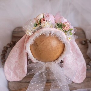 May include: Pink baby bonnet with bunny ears and a floral crown. The bonnet is made of lace and has a tulle bow. The crown is made of pink and white flowers.