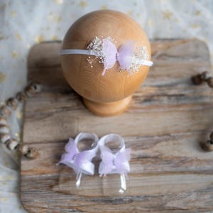 May include: A baby headband and foot sandals set. The headband features a white elastic band with a lavender butterfly and small white flowers. The foot sandals have a similar design. The items are displayed on a wooden tray.