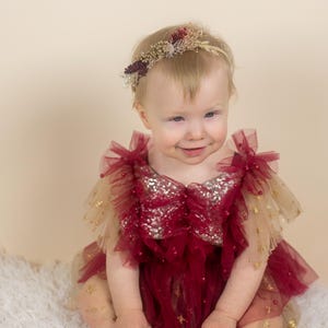 May include: A baby wearing a burgundy and gold tulle dress with a sequined bodice and star accents. The dress has ruffled sleeves and a layered skirt. The baby is also wearing a floral headband. The background is a soft beige.
