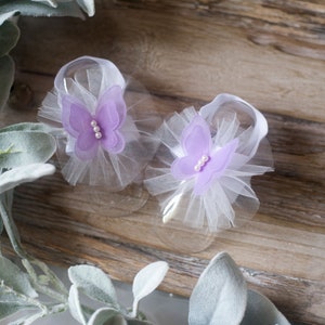 May include: A pair of baby shoes with a white tulle bow and a purple butterfly accent. The butterfly has a pearl center.