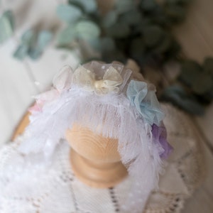 May include: A white lace headband with pastel colored fabric flowers. The headband is decorated with a white bow and has a ruffled edge.