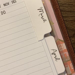 May include: Close-up of a white planner with a tab labelled "March" and another tab labelled "April". The planner has a section labelled "To Buy" and a section with the months November and December.