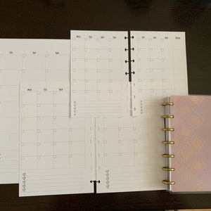 May include: A set of blank calendar pages for a planner, with a pink and gold patterned planner cover. The pages are designed for weekly and monthly planning.