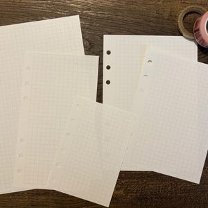 May include: Four white sheets of paper with a grid pattern and holes along the left edge. The sheets are arranged in a staggered pattern, with the largest sheet at the bottom and the smallest sheet at the top.