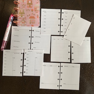 May include: A pink and gold planner with various inserts, including weekly, daily, and monthly layouts. A multi-colored pen rests beside the planner. The planner and inserts are designed for organization and scheduling.