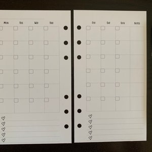Puede incluir: Un inserto de planificador blanco con un dise&ntilde;o de calendario mensual, con casillas para cada d&iacute;a de la semana y espacio para notas. Un bol&iacute;grafo verde azulado descansa junto al planificador. El planificador tiene agujeros perforados para encuadernar.
