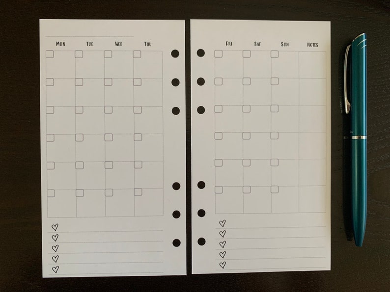Puede incluir: Un inserto de planificador blanco con un dise&ntilde;o de calendario mensual, con casillas para cada d&iacute;a de la semana y espacio para notas. Un bol&iacute;grafo verde azulado descansa junto al planificador. El planificador tiene agujeros perforados para encuadernar.