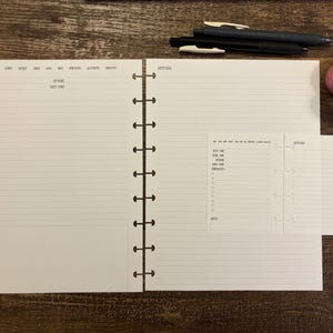 May include: An open planner with lined pages, a smaller insert, and a black pen. Two rolls of decorative tape in pink and gray are also present. The planner is on a wooden surface.