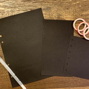 May include: Three black paper sheets with holes along the side, a white pen, and three rolls of pink and red washi tape on a wooden surface.