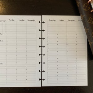 Peut inclure: Un agenda hebdomadaire blanc avec une reliure à spirale noire. L'agenda comporte des colonnes pour chaque jour de la semaine, du lundi au dimanche, et des lignes pour chaque heure de la journée. L'agenda comporte des sections pour "Top 3", "To Do" et "Notes".