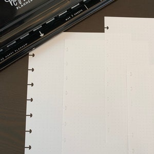 May include: A black Happy Planner punch with the text "The Happy Planner" and "Max 3 Sheets" on it. The punch is next to two white sheets of paper with holes punched along the left side.