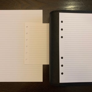 May include: Two white lined sheets of paper with holes on the left side. The smaller sheet is on top of the larger sheet. The larger sheet is on a dark brown surface. A black leather bound notebook with a white lined sheet of paper inside is on the right side of the image.