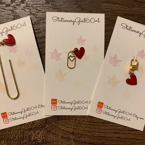 May include: Three cards with the text "Stationery Girl 604" on them. Each card features a different gold-coloured bookmark with a red heart charm. The bookmarks are a paperclip, a large paperclip, and a lobster clasp.