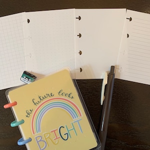 May include: A yellow planner with a rainbow and the text "the future looks bright". The planner has six rings and is open to a blank page. There are four blank pages, one with grid lines, one with dots, and one with lines. A black pen is also pictured.