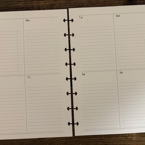 May include: A white paper planner insert with a weekly layout featuring lined sections for each day of the week. The left page includes a Week of header, Monday, and Thursday, while the right page displays Tuesday, Wednesday, Saturday, and Sunday. The paper has mushroom-shaped punch holes along the inner edges.