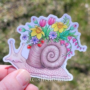 May include: A sticker featuring a whimsical snail with a shell adorned with colorful flowers, including tulips, daffodils, and bleeding hearts. The snail's body is pink, and the shell is a gradient of brown and pink. The text "@wisheaterart" is at the bottom.
