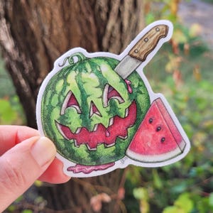 May include: A sticker featuring a green watermelon carved with a jack-o'-lantern face, a knife, and a slice of watermelon. The design has red and pink accents, with a dripping effect. The sticker is held by a hand.
