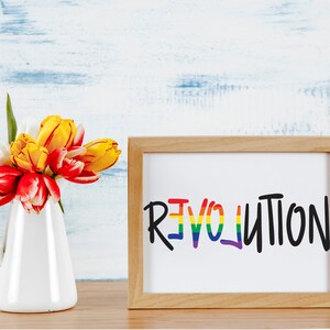 May include: A framed print with the word "REVOLUTION" written in rainbow colors on a white background. The frame is made of light brown wood. A bouquet of yellow and red tulips in a white pitcher is in the foreground.