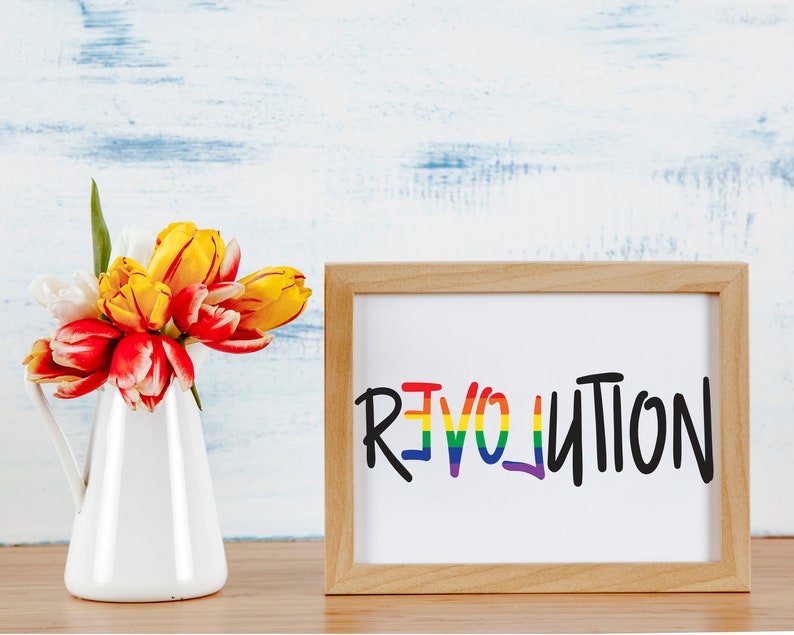 May include: A framed print with the word "REVOLUTION" written in rainbow colors on a white background. The frame is made of light brown wood. A bouquet of yellow and red tulips in a white pitcher is in the foreground.