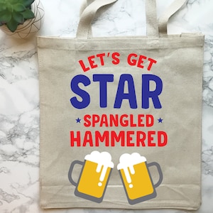 Op de afbeelding: Een witte canvas tote bag met de tekst "Let's Get Star Spangled Hammered" in rode en blauwe letters. Twee cartoon bierbekers met gele vloeistof en wit schuim bevinden zich aan weerszijden van de tekst.