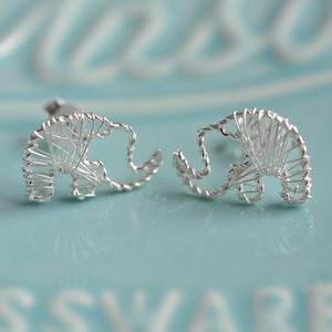May include: A pair of silver-colored elephant-shaped stud earrings. The earrings are crafted from wire, with the elephant's form clearly defined. The earrings are displayed on a light blue surface, creating a contrast with the silver metal.