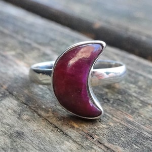May include: A silver ring with a crescent moon shaped gemstone. The gemstone is a deep purple color.