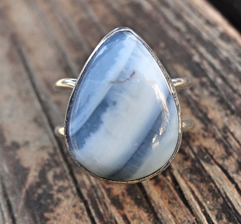 May include: A silver ring featuring a large, teardrop-shaped gemstone with a blue and white striped pattern. The ring has a double-band design, and the stone is set in a silver bezel.