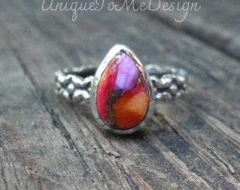 May include: A teardrop-shaped ring with a colorful gemstone set in sterling silver. The gemstone is a mix of pink, orange, and red with gold flecks. The band is a textured silver band.