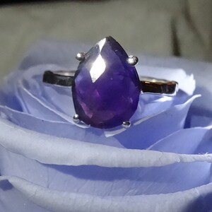 May include: A silver ring featuring a large, pear-shaped amethyst gemstone. The ring is displayed on a blue rose.