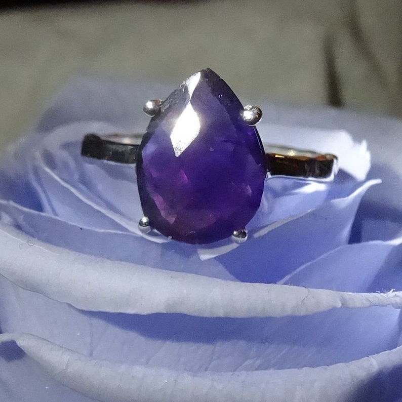 May include: A silver ring featuring a large, pear-shaped amethyst gemstone. The ring is displayed on a blue rose.