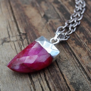 May include: A faceted, deep pink gemstone pendant necklace. The gemstone is capped with a silver-colored metal setting and hangs from a silver chain. The pendant is set against a rustic, wooden background.