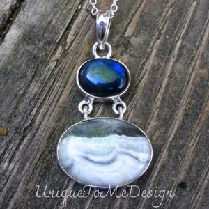 May include: A silver pendant necklace featuring two oval-shaped stones. The top stone is dark blue with green and gold flecks. The bottom stone is white with grey and green swirls. The pendant hangs from a silver chain.