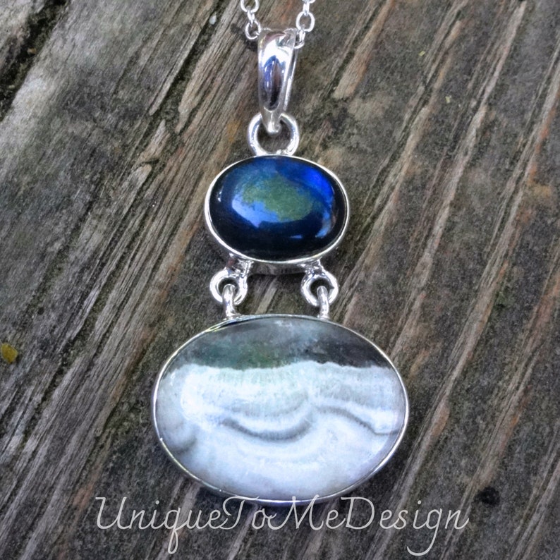 May include: A silver pendant necklace featuring two oval-shaped stones. The top stone is dark blue with green and gold flecks. The bottom stone is white with grey and green swirls. The pendant hangs from a silver chain.