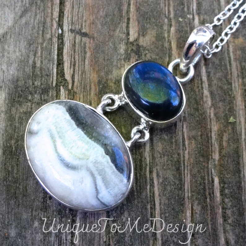 May include: A silver pendant necklace featuring two oval-shaped stones. The top stone is black with a blue iridescent sheen. The bottom stone is white with grey and blue swirls. The necklace is displayed on a wooden surface.
