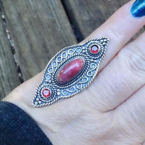 May include: A silver ring featuring an oval pink stone and two smaller red stones. The ring has a detailed design with intricate swirls and lines.