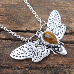 May include: A silver-toned metal necklace featuring a moth-shaped pendant with intricate cut-out patterns and a crescent moon design. The centre of the moth body is set with a smooth, teardrop-shaped brown tiger's eye gemstone with golden bands.