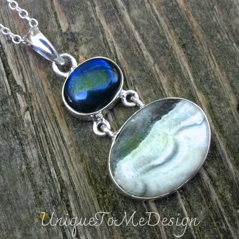 May include: A silver necklace with a pendant featuring two oval-shaped stones. The top stone is a dark blue labradorite with green and blue iridescence. The bottom stone is a white and grey agate with swirling patterns.