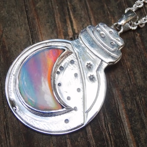 Peut inclure: Un pendentif en argent en forme de bouteille avec un bouchon, orné d'une incrustation de lune croissante en opale irisée. La surface métallique est gravée de petits motifs d'étoiles et de points, suspendu à une chaîne en corde d'argent.
