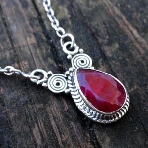 May include: A silver necklace featuring a large, teardrop-shaped, faceted ruby gemstone. The ruby is set in a decorative silver bezel with a twisted rope design. The necklace chain and accents include spiral and swirl designs.