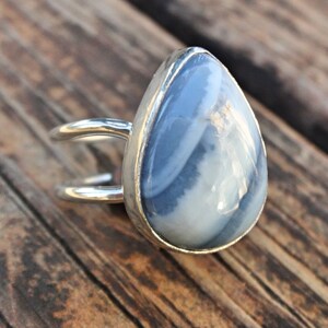 May include: A silver ring featuring a large, teardrop-shaped gemstone with a blue and white marbled pattern. The ring has a double-band design, with the gemstone set in a silver bezel. The ring is displayed on a wooden surface.