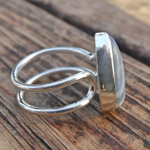 May include: A silver ring with a large, oval, light-colored stone. The ring has a double-band design and is set against a wooden surface. The stone is smooth and polished, reflecting light.