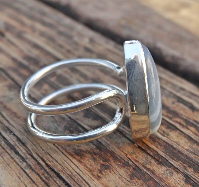 May include: A silver ring with a large, oval, light-colored stone. The ring has a double-band design and is set against a wooden surface. The stone is smooth and polished, reflecting light.