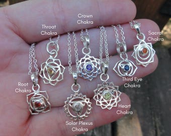 May include: A collection of seven silver-toned chakra necklaces, each with a unique pendant design and a small gemstone. The necklaces are displayed against a blurred background. The text "UniqueToMeDesign" is visible.